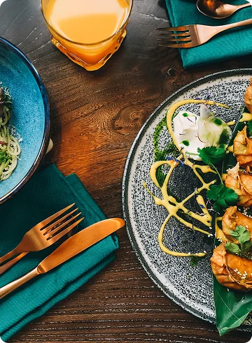 Een professionele foodfoto, genomen van bovenaf (flat lay-perspectief), waarop een verfijnde lunch op een donkere houten tafel met een uitgesproken textuur te zien is. De sfeer is gezellig, modern en smakelijk. Het kleurpalet is gebaseerd op het contrast tussen diepe, donkere houttinten, fel turkoois of donkergroene servetten en rijke goudoranje accenten in het eten en drinken.