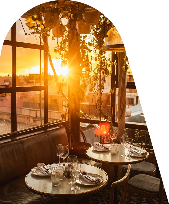 Een foto uit een gezellige fotoshoot in de stijl van een interieurmagazine, genomen in een café met panoramische ramen bij zonsondergang. De sfeer is romantisch, warm en vredig. Het kleurenpalet is gebaseerd op de rijke goud-, honing- en barnsteentinten van de ondergaande zon, die contrasteren met de diepe donkerbruine en zwarte raamkozijnen.
