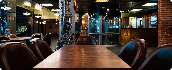 De foto toont het interieur van een modern restaurant of een bar in loftstijl. De ruimte wordt gedomineerd door donkere en warme tinten: donkerbruin, zwart en de kleur van natuurlijk hout, met accenten van geel kunstlicht afkomstig van de plafondlampen. In het midden van het beeld staat een lange, rechthoekige houten tafel op een zwart metalen onderstel, omringd door diepe, donkerbruine leren fauteuils.